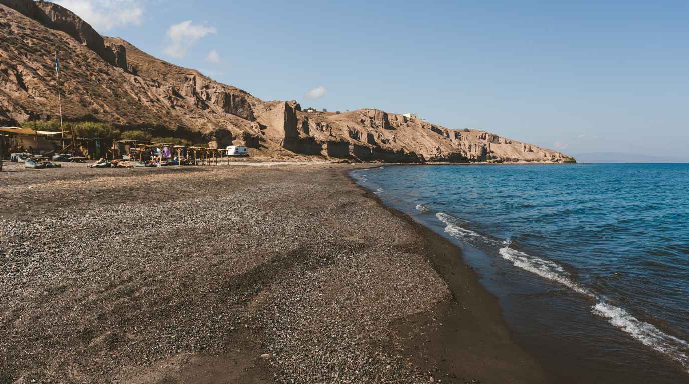 Dark grey volcanic pebbles and clear turquoise water along the quiet shore at Exo Gialos beach, Santorini