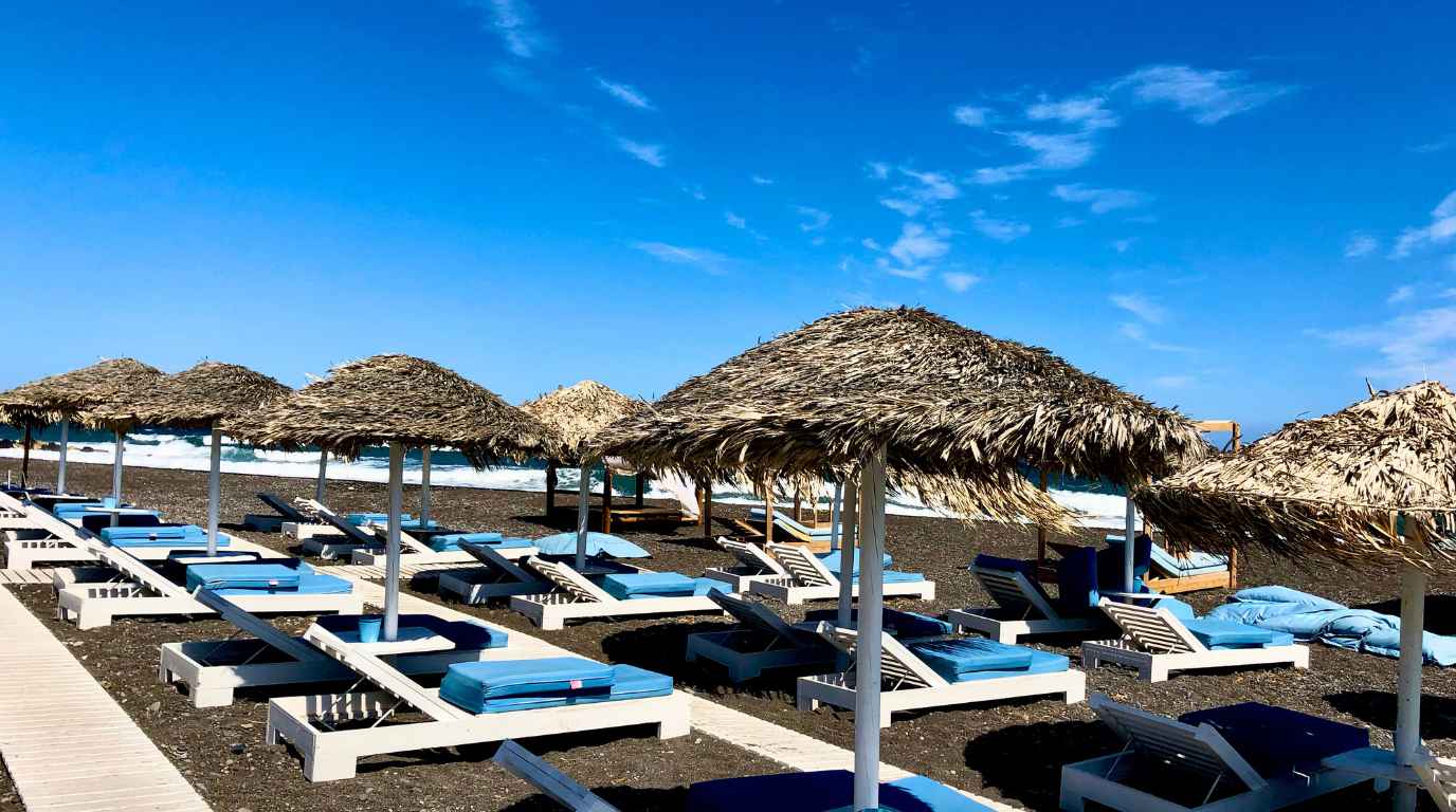 Thatched umbrellas and blue-cushioned sunbeds on the black sand at Perissa beach, Santorini
